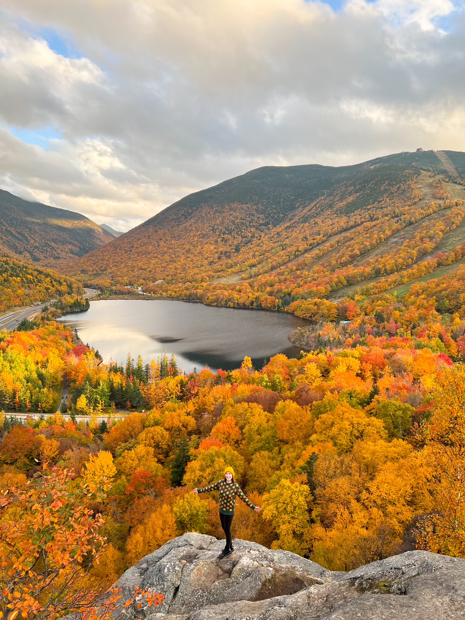 A women in a yellow beanie stands on the edge of a cliff, overlooking the fall foliage of New Hampshire. The fall colors of the leaves on her shirt match the tree leaves behind her. No wonder New Hampshire is one of the prettiest states in the USA! 