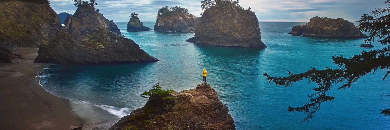 A women in a yellow jacket stands on a cliff and overlooks the dark landscape with the bright blue ocean in Oregon, one of the prettiest states in the USA!