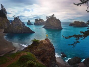 A women in a yellow jacket stands on a cliff and overlooks the dark landscape with the bright blue ocean in Oregon, one of the prettiest states in the USA!