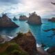 A women in a yellow jacket stands on a cliff and overlooks the dark landscape with the bright blue ocean in Oregon, one of the prettiest states in the USA!