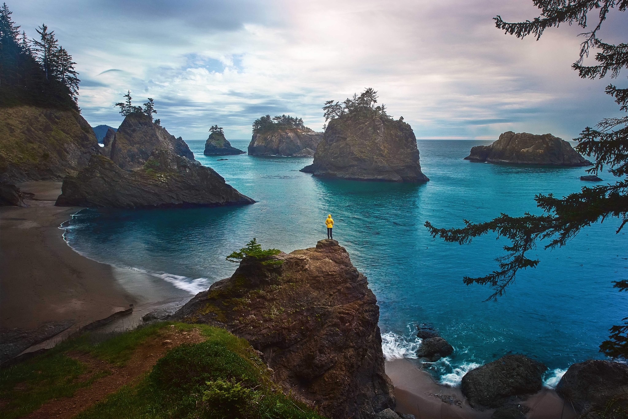 A women in a bright yellow rain jacket is a tiny spec on a rock formation as she overlooks the sea, which is turquoise, in Oregon. 