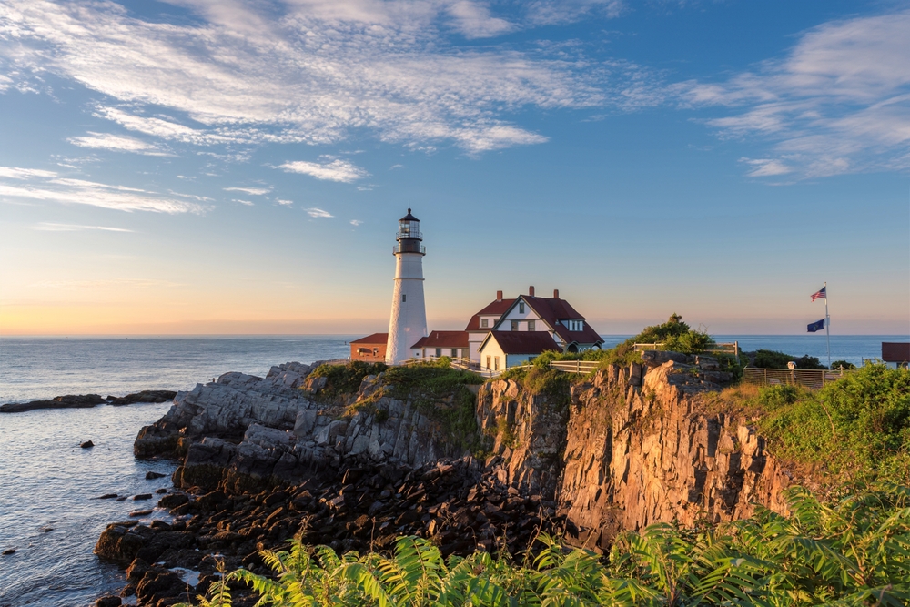 The cliffs and coast of Maine are stunning and make it one of the prettiest states in the USA: this photo shows a lighthouse and a town red and white in color overlooking the rocky Cliffside during sunset. The sun lightens the green grass and blue waters.  