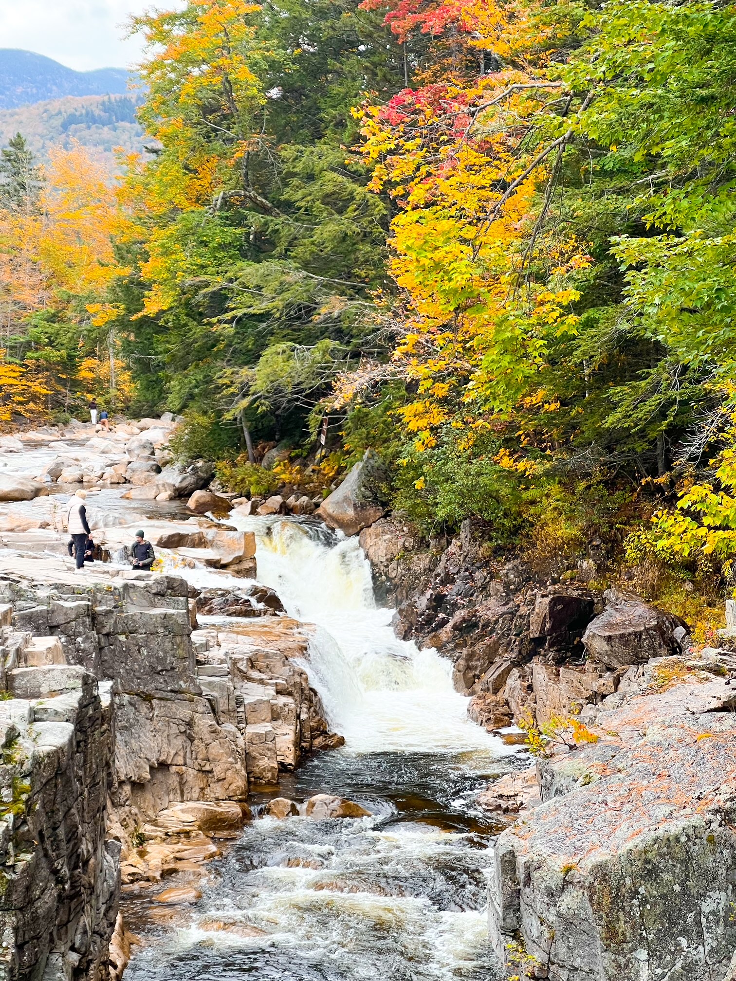 A small waterfall cascades down stone rocks as people climb by it and the green leaves slowly turn red and yellow. 