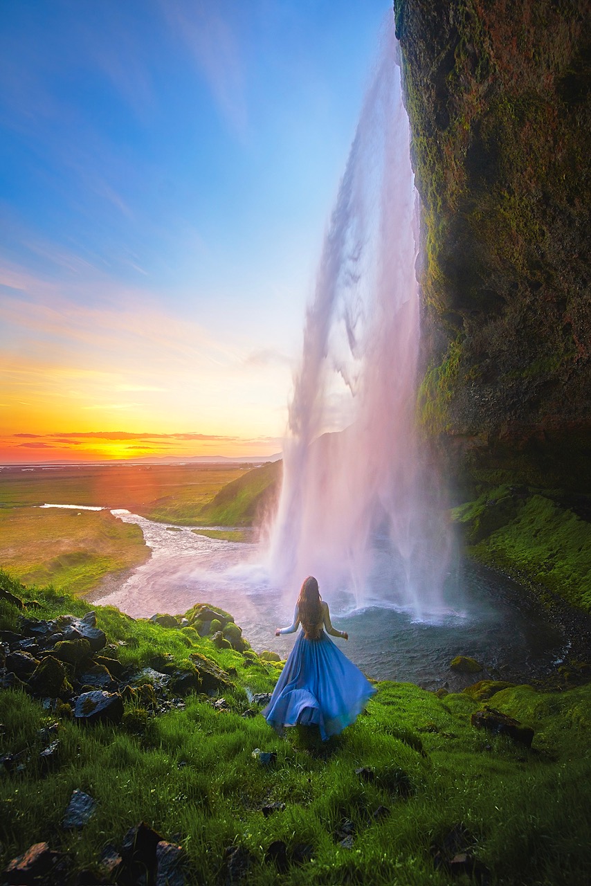 Me in a blue dress looking at one of the many beautiful waterfalls in Iceland. This waterfall is cascading over a cliff into a pond, surrounded by green grass at sunset. 