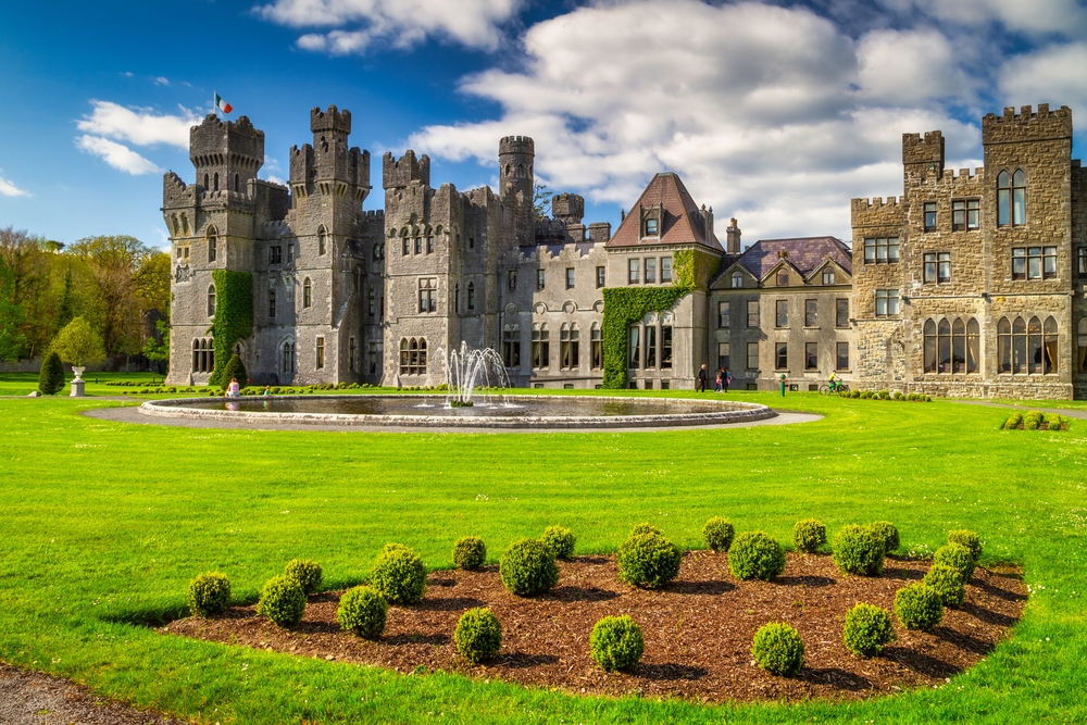 Ireland's stunning Ashford Castle with a fountain in front, surrounded by green lawns and topiaries, and blue skies with scattered clouds. 