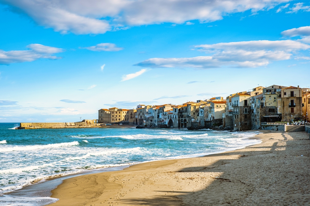 ocean on the left and a sandy beach on the right, in the back right of the photo there are buildings on a bright sunny day 