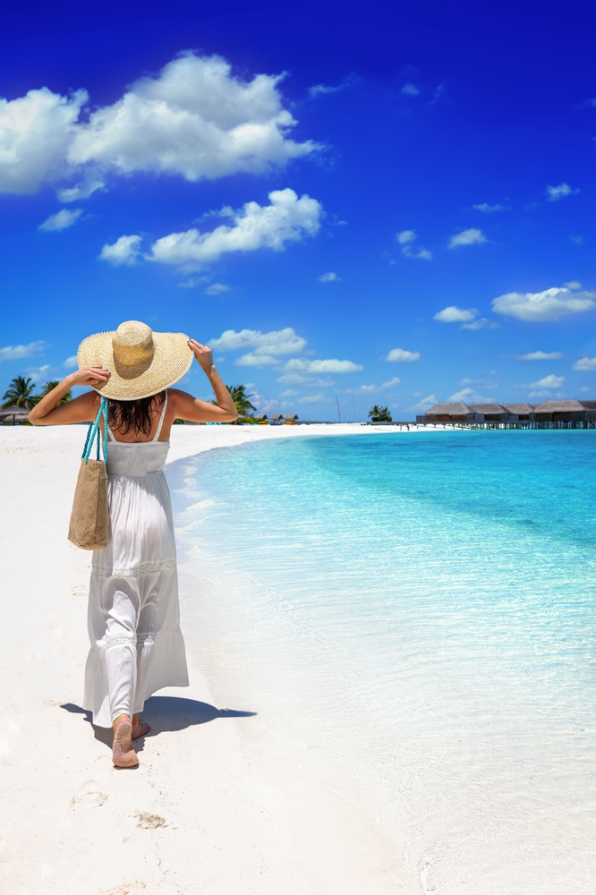 Woman in a white dress and hat walks down a tropical paradise beach in the Maldives islands.