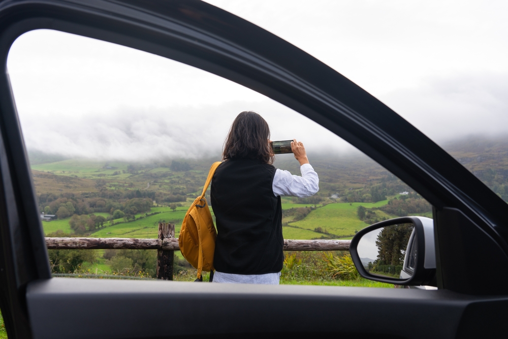 A women has pulled off to the side of the road to take a picture of the fog rolling over the greens in Ireland: her car window frames the photo of her standing outside the car itself.