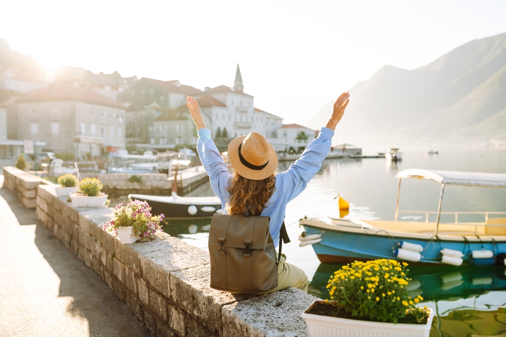 A young woman in a hat sits on the edge of a river, overlooking boats and a city with her hands up in the air, knowing she is aware of the mistakes to avoid in Europe.