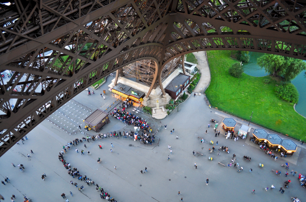 An overhead photo shows the bottom of the Eiffel Tower and the line that snakes around the grounds as people wait in line for a ticket to go up to the top. 