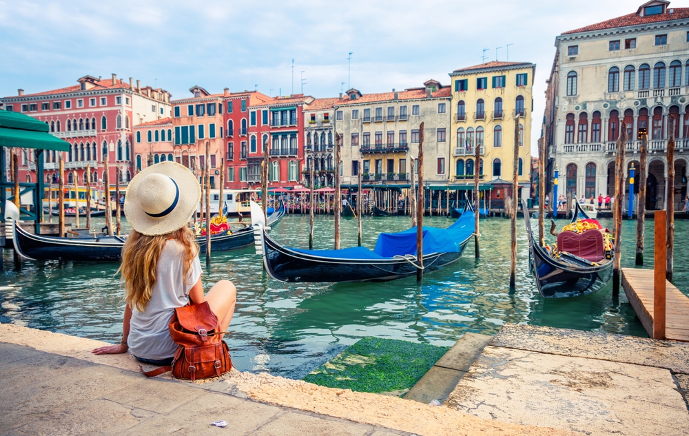 Another young woman in a sun hat sits on the edge of the river, looking across the river at colorful buildings in Venice, as boats rock in the water. 