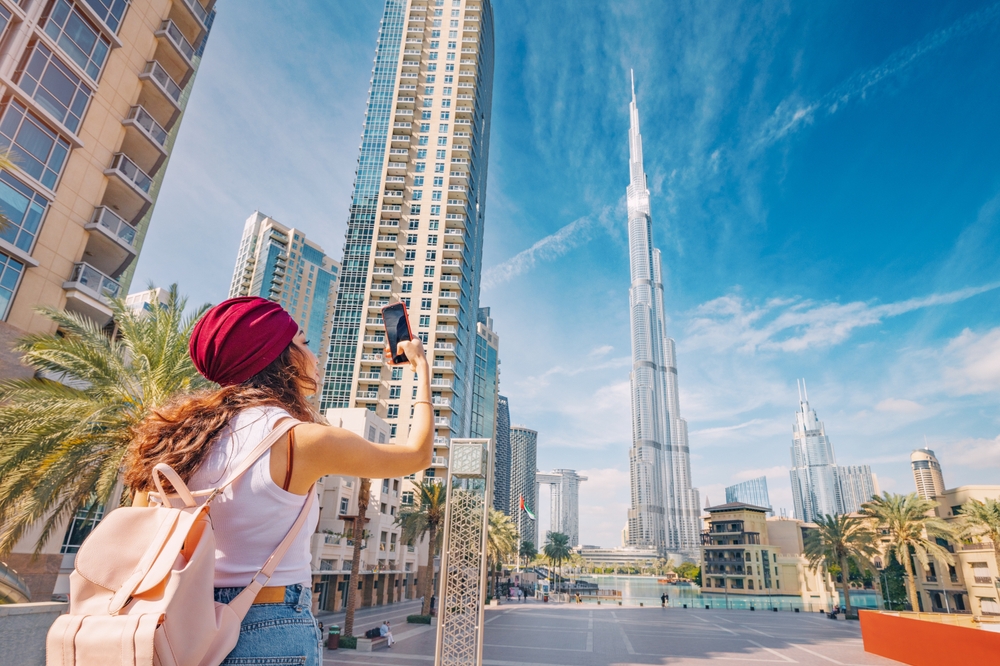 A women with a red head covering and pink backpack points her phone up at a tall building, snapping photos, enjoying her free cell data with an international data plan. 