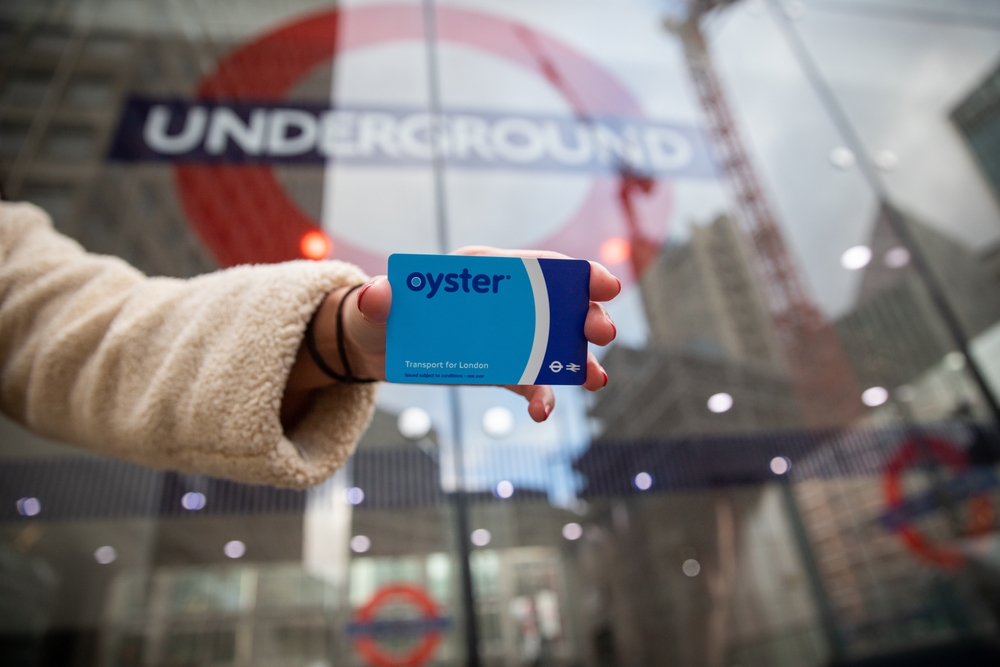 Hand of a girl holding an Oyster card outside the entrance of the underground in the background. The card allows access to the underground in London. This is a mistale to avoid in London. 