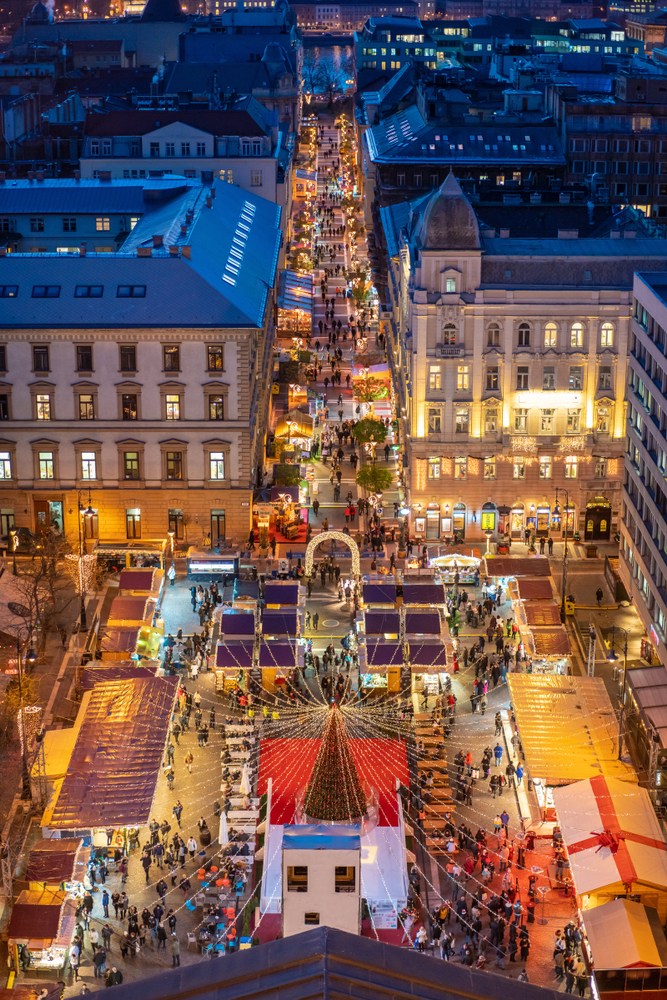 An arial shot of Budapest shows the city at night time during Christmas during their market: the warm glow of lights highlights their town square as people walk and from stall to stall.