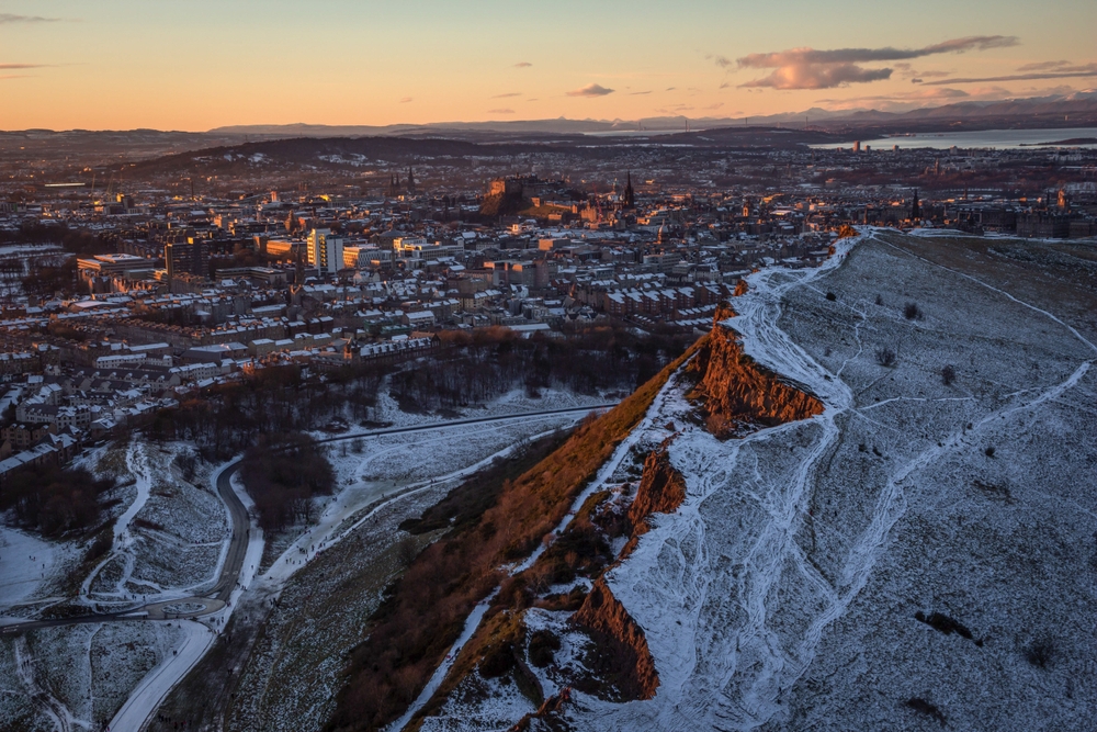 A view from a hike in Edinburgh shows the city from an angle as the snow has covered rooftops and hiking paths. The golden hour is light in the sky. 