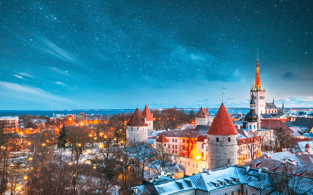 A snowy and starry night at Tallinn shows a blue sky, warm gold lights, and fresh snow falling on the red roofs of the city. 