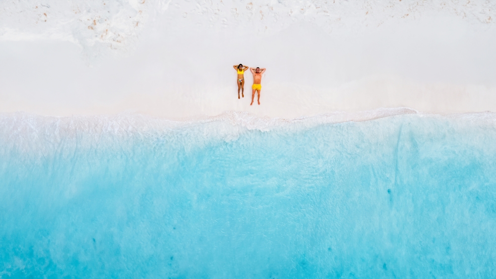 a couple laying on the white sand while a drone takes an Ariel shot of the ocean and beach 