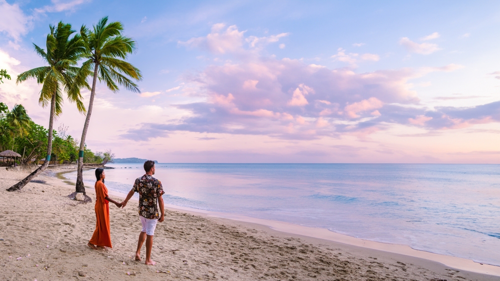 a couple holding hands standing on the beach in St, Lucia at sunset with two palm trees on the beach with cotton candy sky it is one of the most romantic Caribbean honeymoon destinations