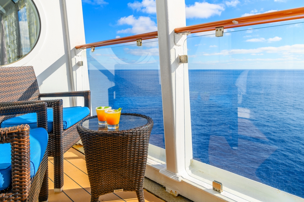 On the balcony of a cruise, blue water is the backdrop with two rinks sitting on the tiny table and chairs of the suite. 