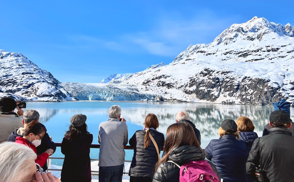 Guests gather at the cusps of a cruise ship, overlooking the icy water of Alaska and the snowcapped mountains, ready to get off and explore. 