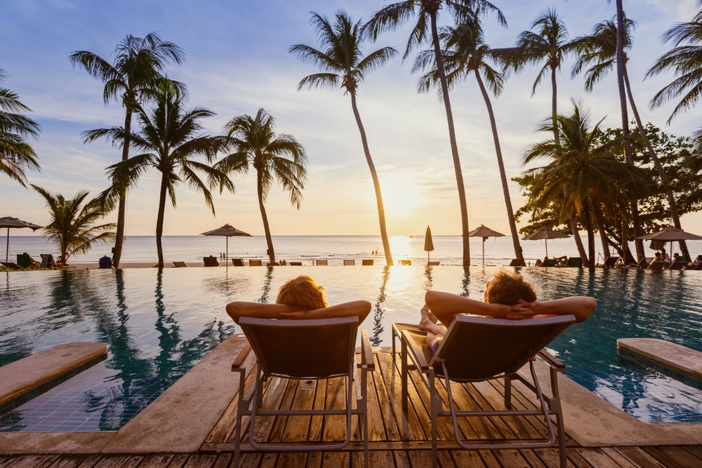 When looking at  all inclusives vs cruises, consider what is best for you and your vacation: this couple lounges on private chairs at a private pool on a private beach at sunset with palm trees nearby. 