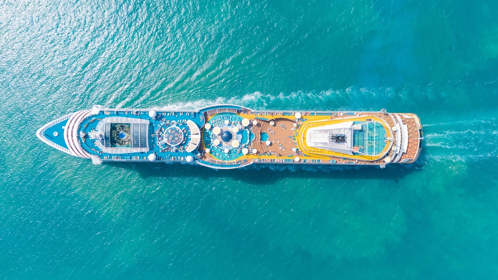 An arial shot of a cruse ship shows it cutting through the water, people on deck, pools, tables, umbrellas and more. 