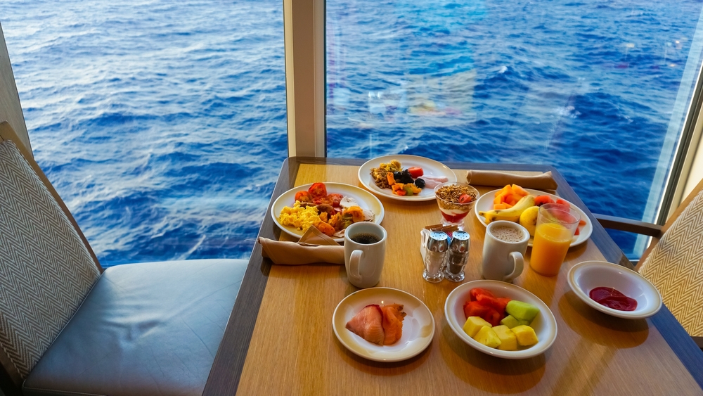 Breakfast sits on a small table: fresh fruit, eggs, and coffee overlook the blue water at a window view on this cruise ship. 