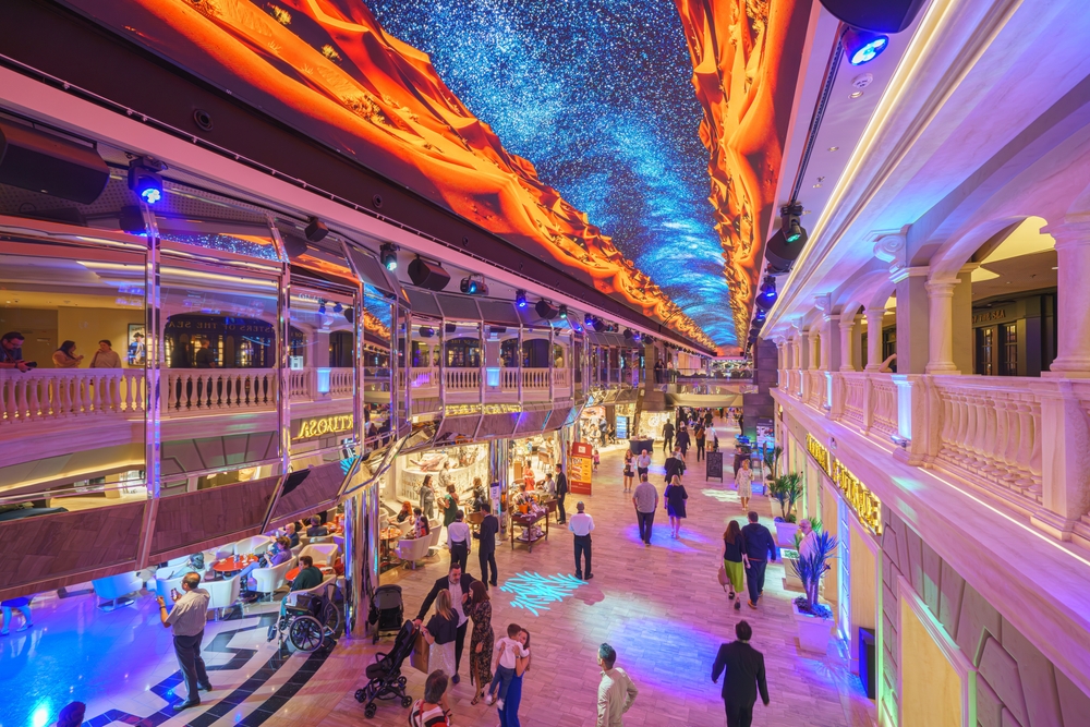 The interior of a cruise ship shows people walking through gate endless options of food, shops and more. The ceiling glows with stars and fire-like imagery. 