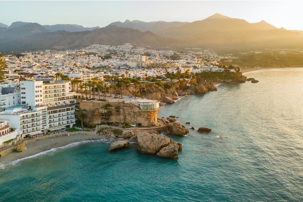 ocean on the right, land on the left with many white buildings, mountains in the background 