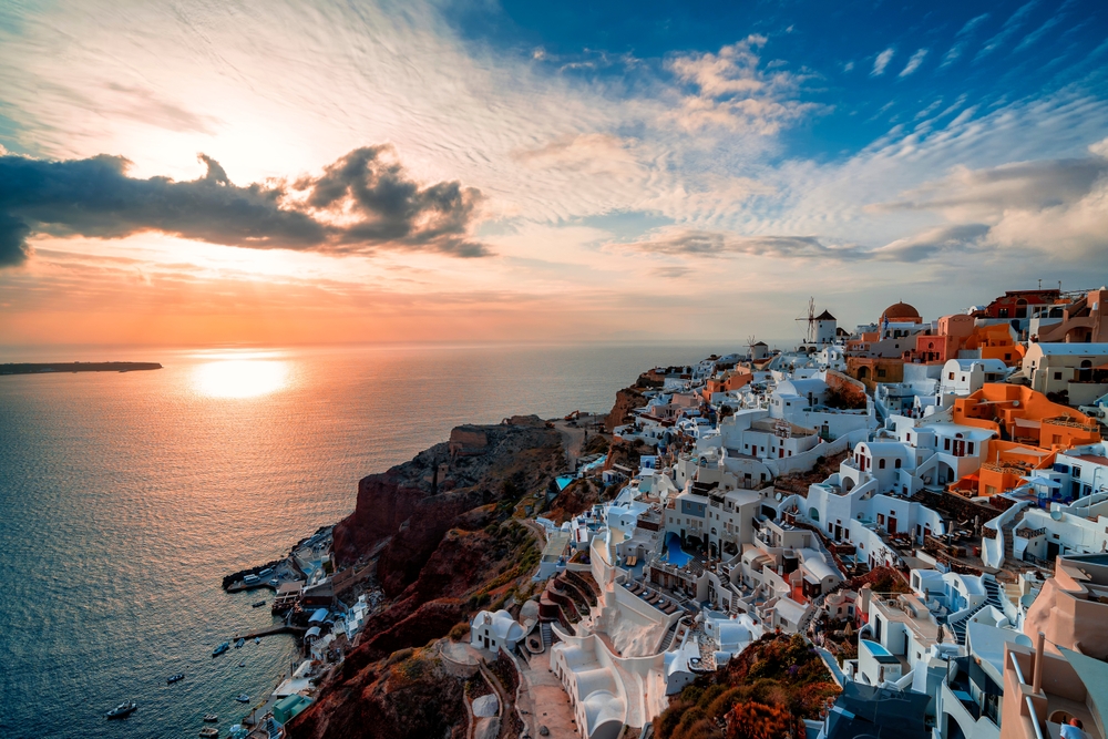 buildings up a hill at the rise in Santorini, Greece buildings are on the right water is on the left. There are clouds in the sky.