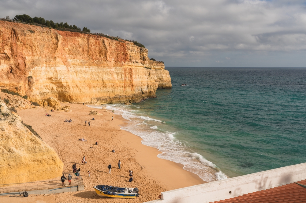 large rock cliff in background on the left, and the ocean is on the right. there are people standing on a sandy beach by the water