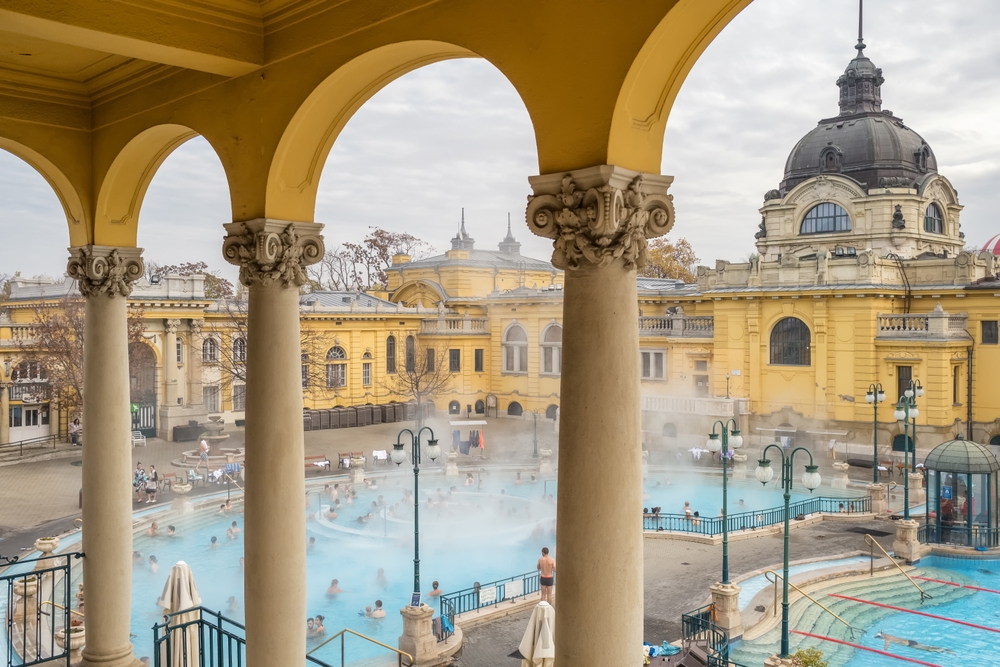 One of the places to visit in Europe in January Budapest, hungry in the foreground there are archways with columns in the middle of the image. You can see a thermal bath with people in it in the background there is a building with a dome top.