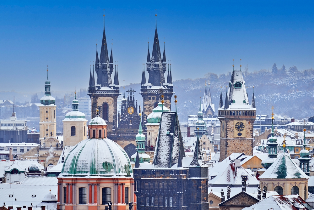 The two spires in the middle of the image in the foreground there is a domed building. All of the buildings are covered in snow, and in the background, there are snow covered trees.