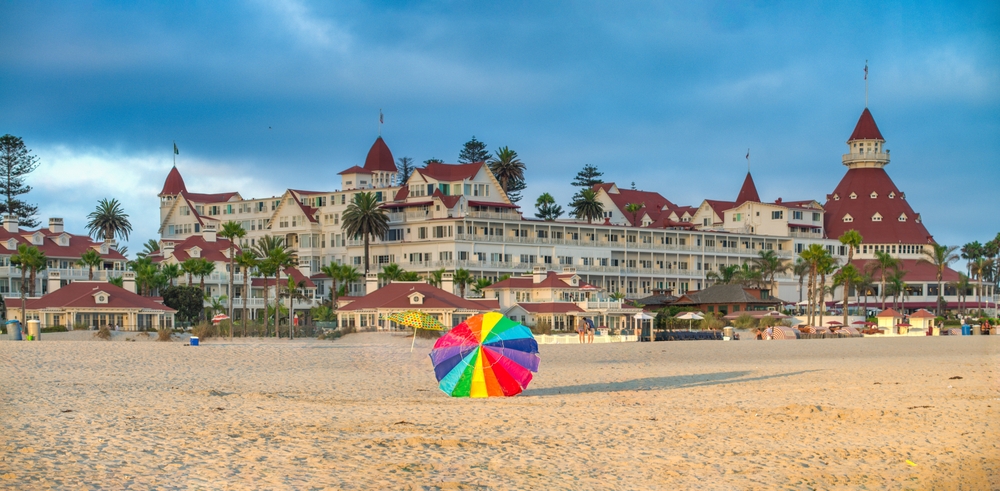 the beach in Coronado, California in the foreground with a rainbow colored umbrella lying on the sand and the famous Hotel Del Coronado in the background