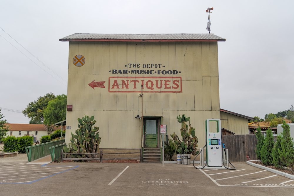 an old, brown antique shop called The Depot advertising a bar, music, and food, with prickly pear cacti and a parking lot in font of the building