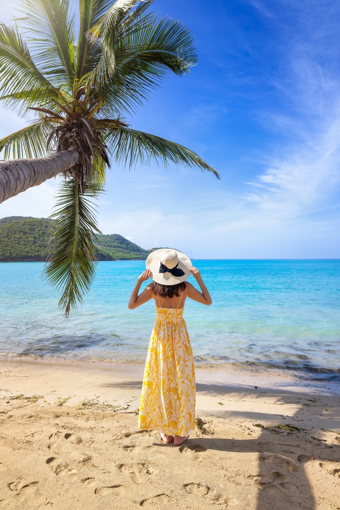 A woman stands on a sandy beach, facing the turquoise ocean while wearing a yellow floral sundress and a wide-brimmed straw hat with a black ribbon. A tall palm tree leans overhead, and the sun shines brightly in the clear blue sky, creating an idyllic tropical setting perfect for a Caribbean vacation. The outfit is light and breezy, ideal for enjoying a warm day by the sea.