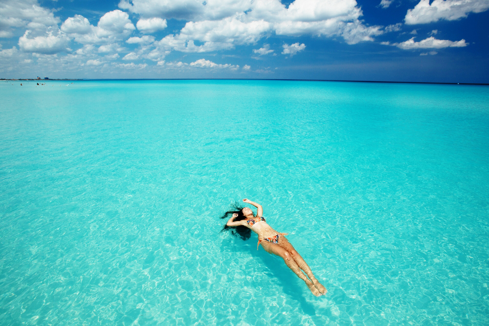 A woman floats peacefully in crystal-clear turquoise water, wearing a patterned bikini. The vast, calm ocean stretches out under a bright blue sky with soft clouds, creating an idyllic and serene Caribbean vacation scene. The image captures a moment of pure relaxation and escape.