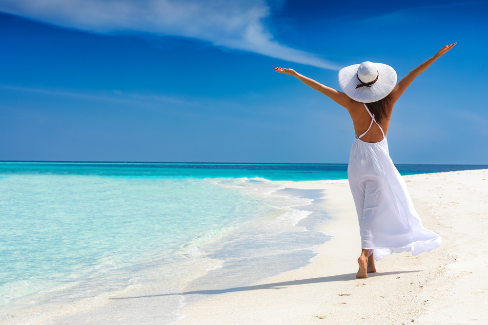 A woman raises her arms in joy as she walks along a white sandy beach, wearing a flowing white halter maxi dress and a wide-brimmed straw hat. The clear turquoise water and blue sky enhance the serene, carefree feeling of this beach vacation look.