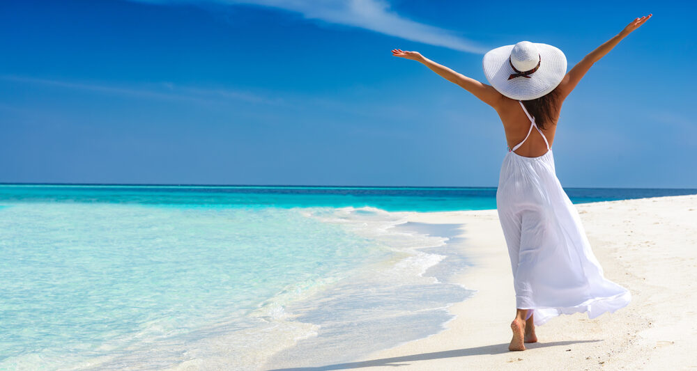 woman standing in white dress with her hands in the air as she walks along the beach