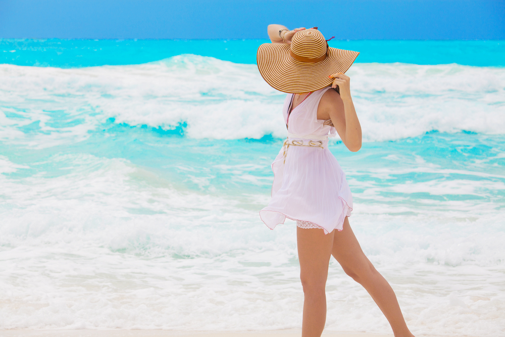 A woman stands by the edge of the ocean, wearing a flowy white dress and a large, striped sun hat. She holds the brim of her hat as the wind blows, with bright turquoise waters in the background. The outfit and setting reflect a breezy, sun-soaked day, perfect for a Caribbean vacation.