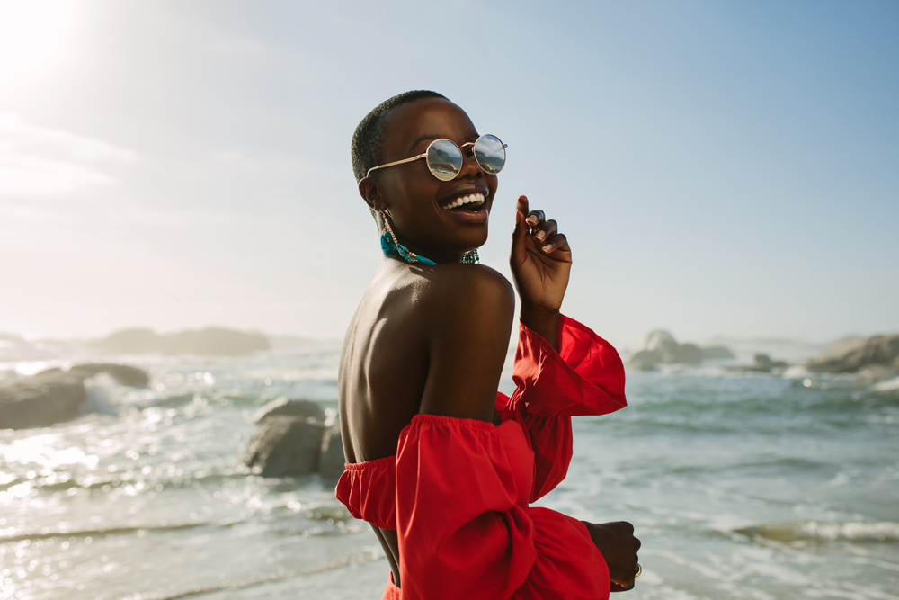 A woman stands on a beach with the ocean in the background, smiling and wearing a vibrant off-the-shoulder red top, paired with sunglasses and statement earrings. The sunny, tropical setting and her carefree expression capture the essence of a warm and relaxing Caribbean vacation. This outfit is perfect for a stylish yet comfortable beach look.