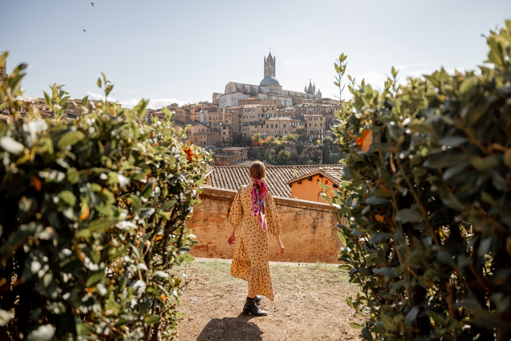 woman walking in a polka dot outfit in italy walking through tuscany vineyards with the town of siena in the background