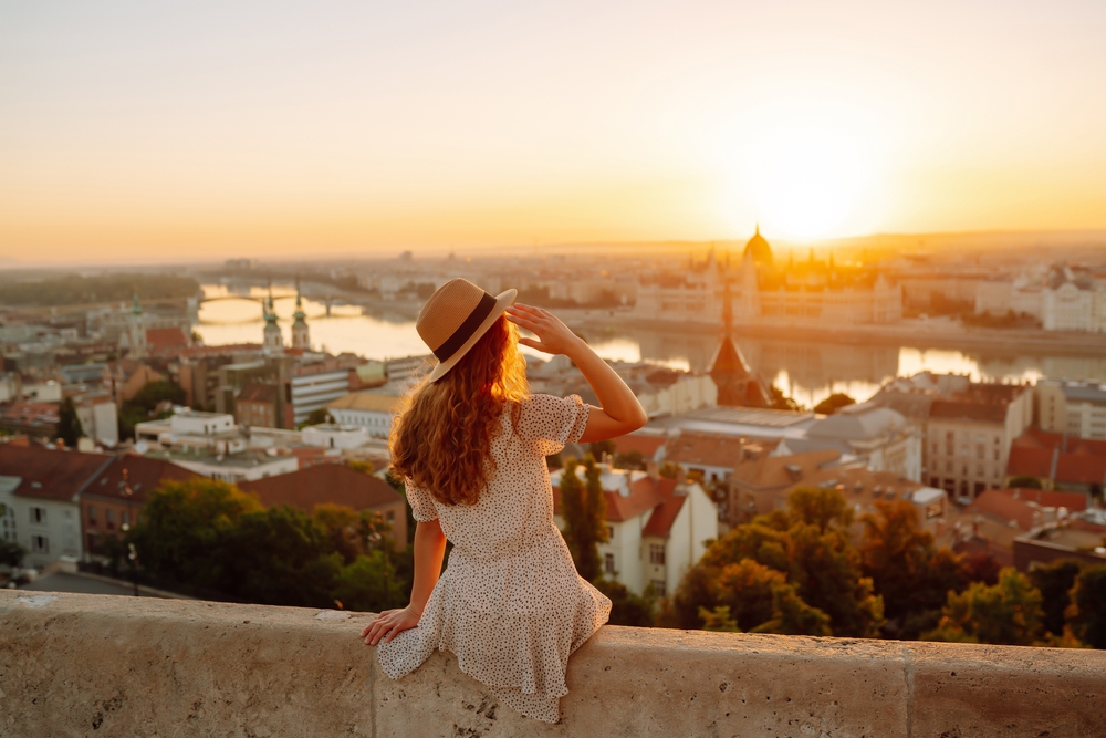 A woman in a light summer dress and straw hat sits on a stone ledge overlooking a cityscape at sunset, gazing towards the distant skyline with a river and iconic buildings bathed in golden light.