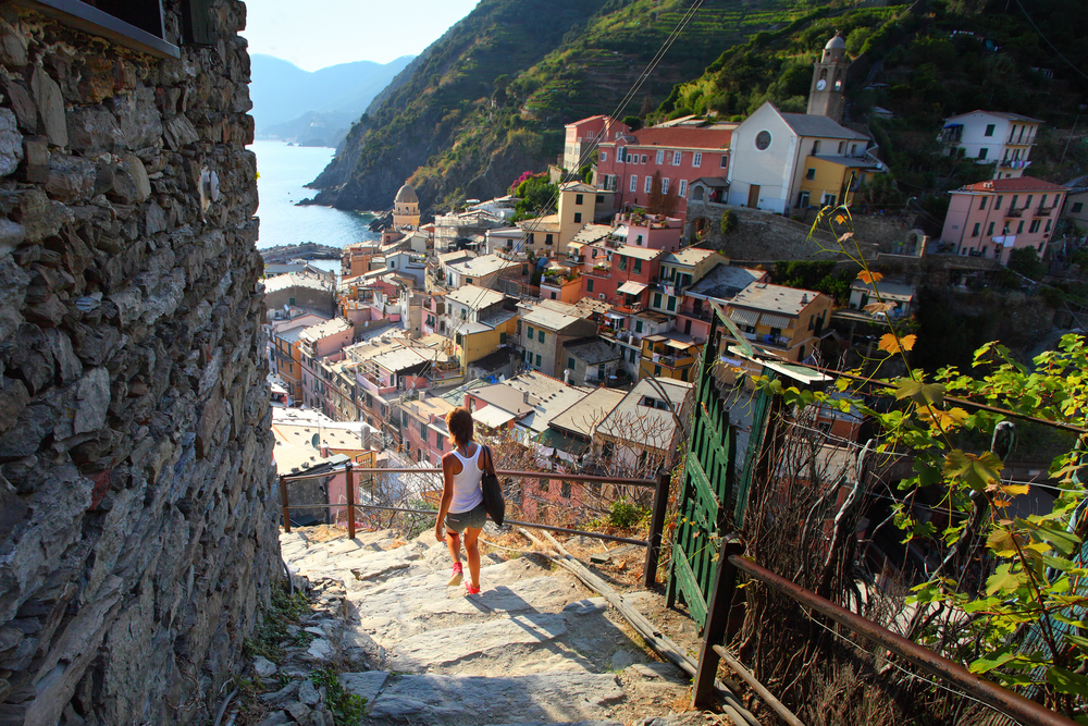 A woman descends a steep staircase in a quaint village, wearing casual summer attire. Below her, colorful houses and buildings overlook a scenic coastline with mountains in the distance.