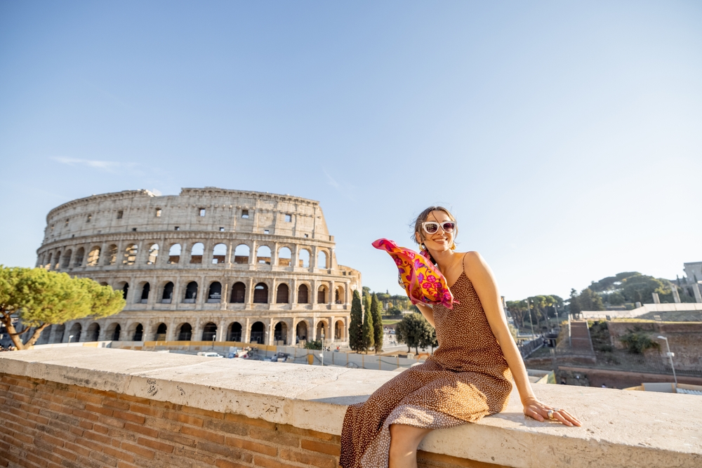 A woman wearing a stylish dress, sunglasses, and a colorful scarf sits on a ledge with the Colosseum in Rome as a backdrop. She smiles at the camera, with a sunny, clear blue sky overhead.