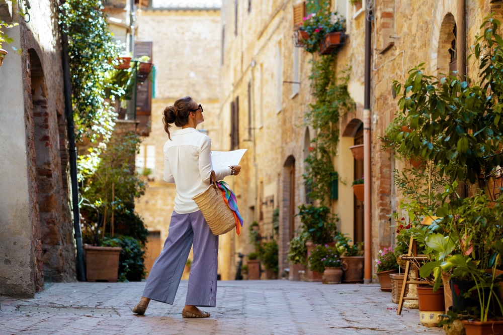 A woman exploring a narrow, picturesque street in a historic town, wearing a white shirt and purple pants, holding a map and a straw bag. The stone walls around her are adorned with potted plants and greenery.