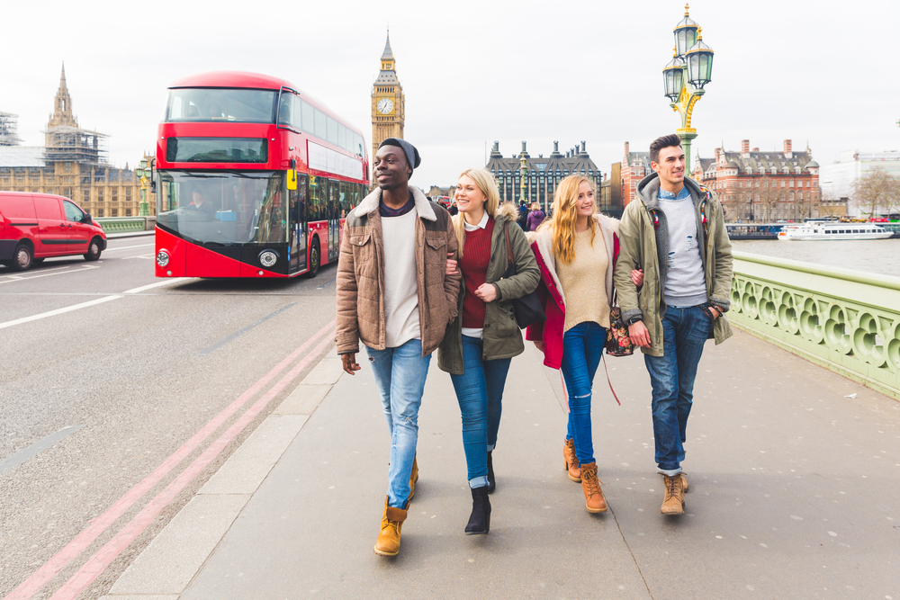 a group of four friends walking across a bridge in London with Big Ben and the Houses of Parliament in the background. They are dressed in casual winter outfits, including jackets, sweaters, scarves, and boots. A red double-decker bus drives by, adding to the iconic London setting. The scene suggests a lively, cold-weather day in December, ideal for showcasing winter fashion in London.