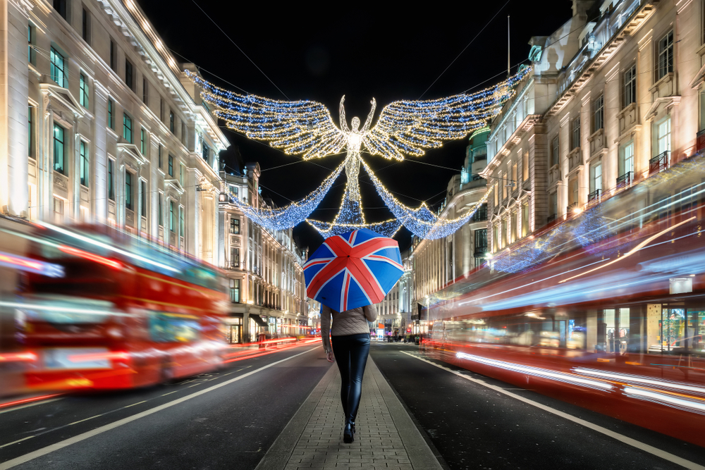  a person walking down a brightly lit street at night, holding an umbrella featuring the Union Jack (UK flag). The street is decorated with festive Christmas lights, including a large angel figure made of twinkling lights overhead. Red double-decker buses are seen in motion, adding to the vibrant, iconic London atmosphere. The scene captures the festive energy of London during the holiday season.