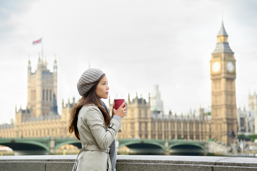 a woman standing near the Thames River in London, holding a takeaway coffee cup. She is dressed in a light trench coat and a grey knit beanie, perfect for a chilly day. In the background, the iconic Big Ben and the Houses of Parliament are visible, adding to the quintessential London scene. The photo evokes a calm, reflective moment during a winter day in the city.