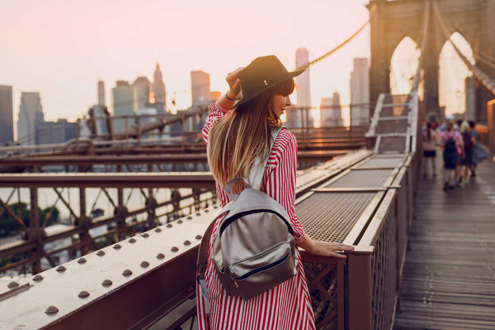 A woman in a striped dress and black hat stands on a bridge, likely the Brooklyn Bridge, during sunset. She has a silver backpack and is gazing toward the city skyline, with the sun casting a warm glow over the scene. The perspective captures a serene, urban moment.